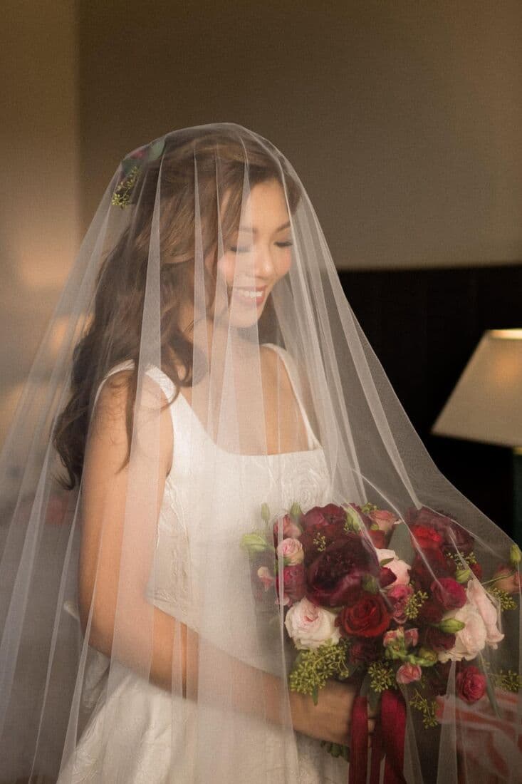 A bride beneath her veil holding a deep red bouquet