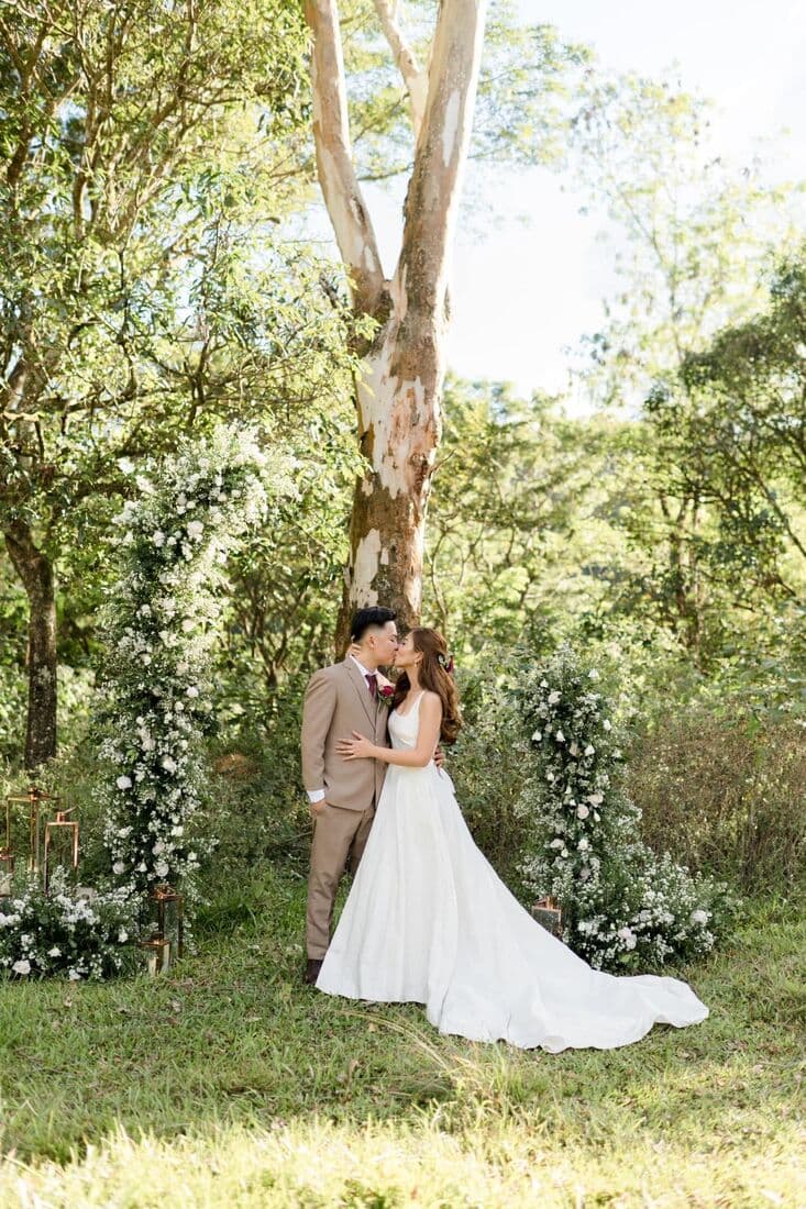 A bride beside a floral arch under tall trees